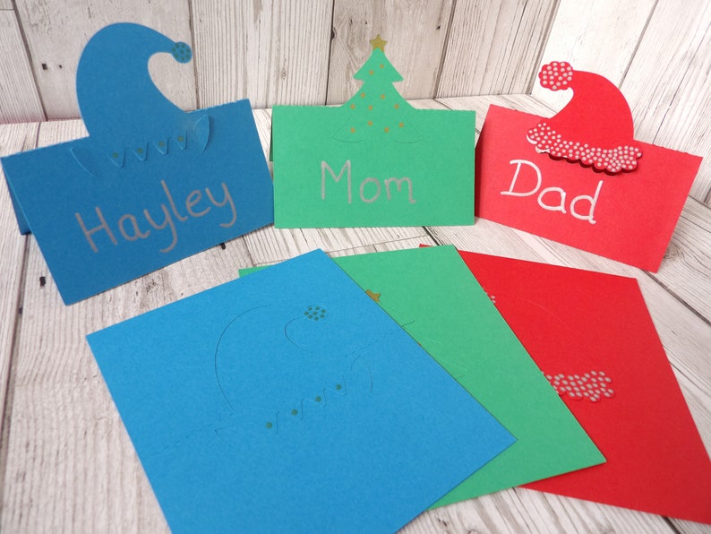 A blue place card with an elf hat cut out of the top, a green place card with a Christmas tree cut out of the top and a red place card with a Santa hat cut out of the top. The 3 place cards are also shown flat in front.