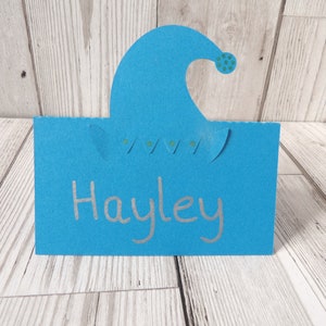 A blue place card with an elf hat cut out of the top. The hat has metallic green dots on the bobble and trim. Hayley is written on the card as an example of it in use.