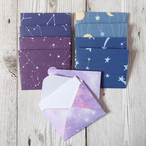 May include: A set of colourful envelopes with a constellation pattern. The envelopes are in shades of purple, blue, and grey. The envelopes are arranged in a stack with a single envelope in front of the stack. The single envelope is a light purple with a galaxy pattern.