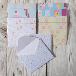 May include: A selection of patterned envelopes featuring cupcakes, balloons, ice cream, and donuts. One envelope is open, showing a white interior. The envelopes are arranged on a light wood-grain surface. The designs are in pastel colours.