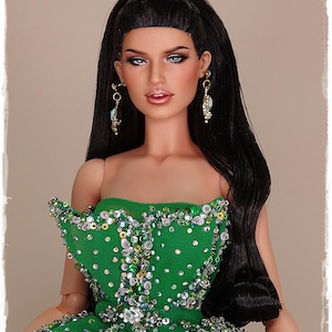 May include: A fashion doll with long black hair and striking makeup. She is dressed in a strapless green gown adorned with sequins and beads. The doll has dangling earrings against a neutral backdrop.