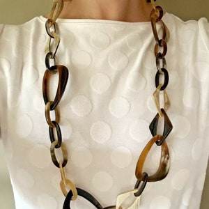 May include: A long, chunky necklace featuring a variety of horn-shaped links in shades of brown, tan, and black. The necklace is composed of oval, teardrop, and diamond-shaped links. The necklace is worn over a white, textured top.
