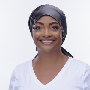 May include: A woman with dark skin is smiling and wearing a black satin headscarf. She is wearing a white v-neck t-shirt.