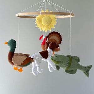 May include: A hanging mobile featuring a circular wooden frame with white strings suspending a yellow felt sun, a mallard duck, a brown and white spotted dog, a turkey with brown feathers, and a green fish, all crafted from felt fabric.