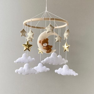 May include: A wooden baby mobile with a crescent moon, a teddy bear, and gold glitter stars hanging from white felt clouds.
