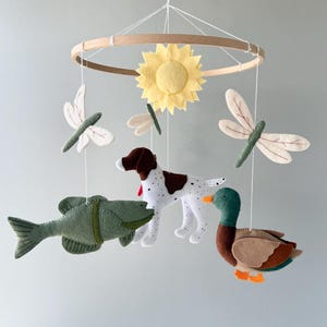 May include: A hanging mobile featuring a circular wooden frame with felt figures suspended by white strings. The figures include a yellow sunflower, two white and green dragonflies, a green fish, a white and brown spotted dog, and a mallard duck.