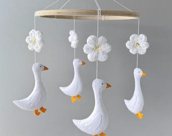 Op de afbeelding: Een baby mobile met witte vilten ganzen met oranje voeten en snavels, en witte bloemaccenten. De ganzen en bloemen hangen aan witte touwtjes aan een houten ring. De mobile hangt tegen een lichtgrijze achtergrond.