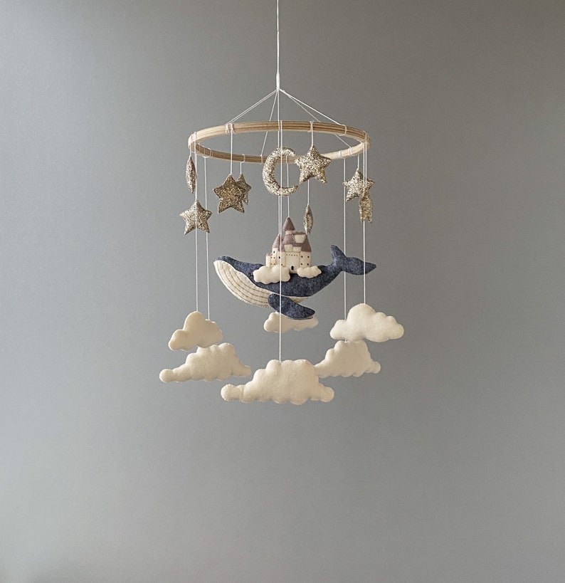 Puede incluir: Un m&oacute;vil de beb&eacute; de madera con una ballena azul, nubes y estrellas doradas. La ballena sostiene una peque&ntilde;a casa.
