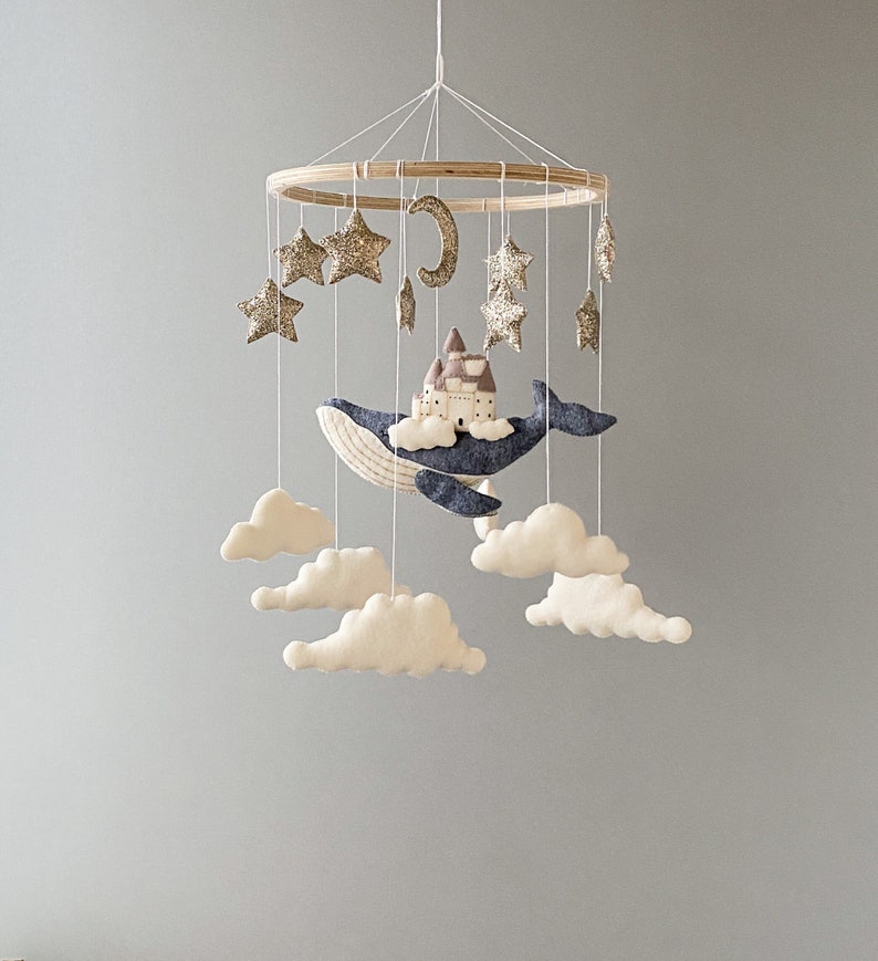 Puede incluir: Un m&oacute;vil de beb&eacute; de madera con una ballena azul, nubes blancas y estrellas doradas con purpurina. La ballena lleva una peque&ntilde;a casa marr&oacute;n con un techo blanco.