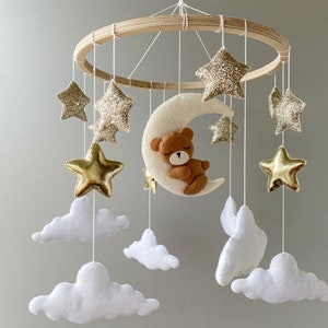 May include: A wooden baby mobile with a crescent moon, stars, and clouds. The moon has a brown teddy bear sleeping on it. The stars are gold and glitter, and the clouds are white.