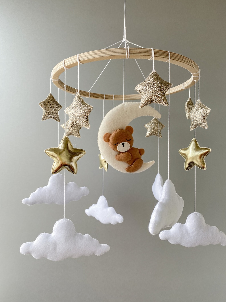 May include: A wooden baby mobile with a crescent moon, stars, and clouds. The moon has a brown teddy bear sleeping on it. The stars are gold and glitter, and the clouds are white.