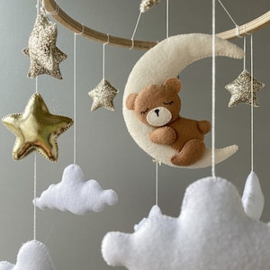 May include: A wooden baby mobile with a crescent moon, stars, and clouds. The moon has a brown teddy bear sleeping on it. The stars are gold and glitter, and the clouds are white.