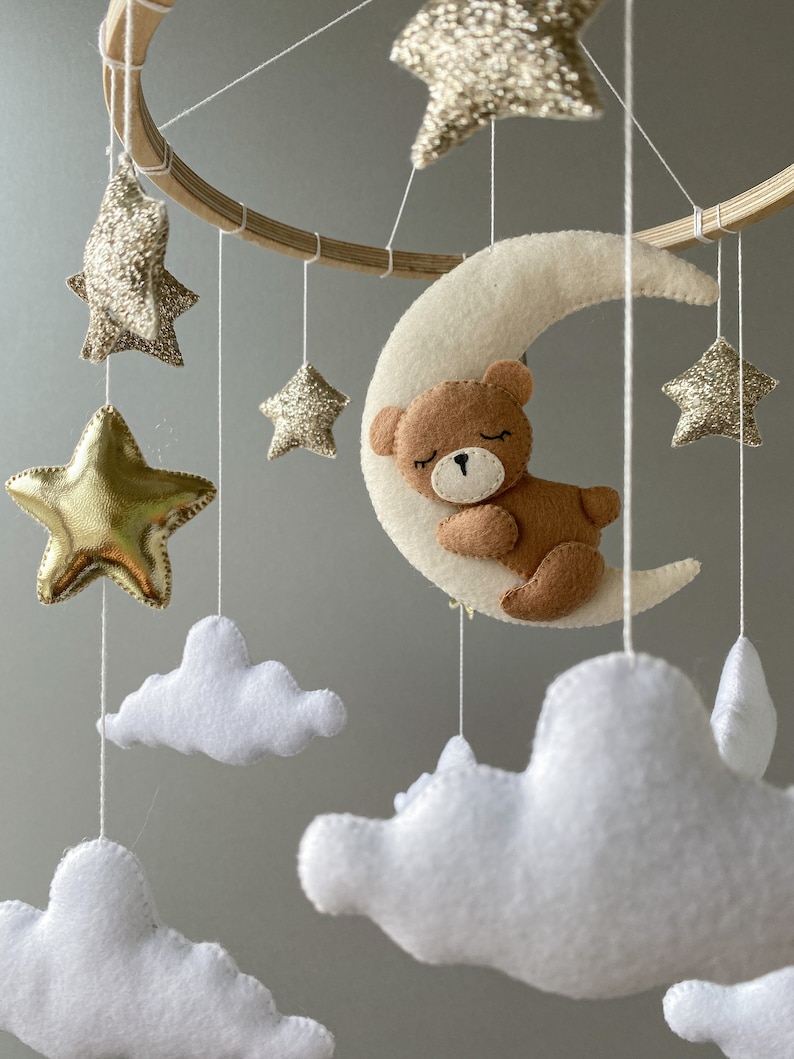 May include: A wooden baby mobile with a crescent moon, stars, and clouds. The moon has a brown teddy bear sleeping on it. The stars are gold and glitter, and the clouds are white.