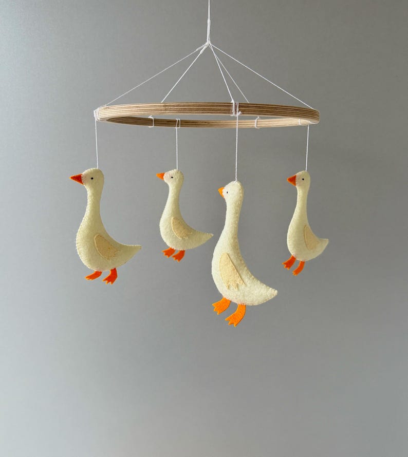 May include: A wooden ring mobile with four felt geese hanging from it. The geese are yellow with orange beaks and feet.