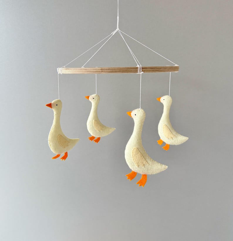 May include: A wooden mobile with four felt geese hanging from it. The geese are white with orange beaks and feet. The mobile is suspended from a white string.