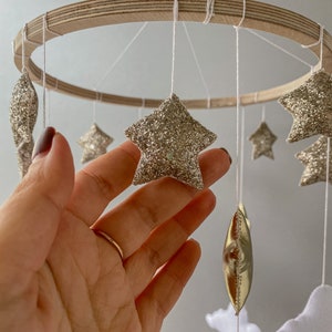 May include: A wooden baby mobile with gold glitter stars, gold foil crescent moons, and white clouds. The mobile is hanging from a wooden ring.