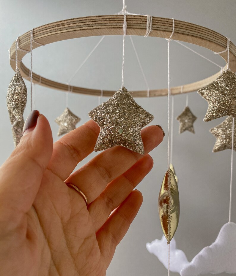 May include: A wooden baby mobile with gold glitter stars, gold foil crescent moons, and white clouds. The mobile is hanging from a wooden ring.
