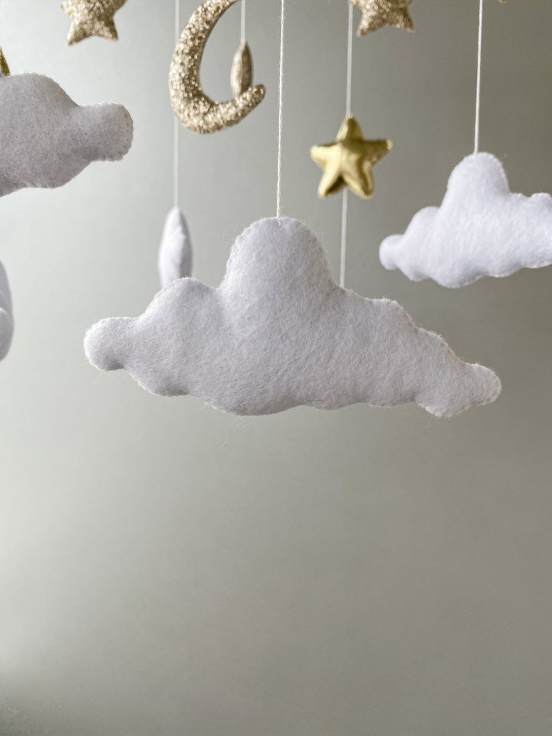 May include: A white felt cloud mobile with gold glitter stars and a crescent moon hanging from a white string.