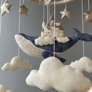Puede incluir: Un m&oacute;vil para beb&eacute; con una ballena azul, nubes blancas y estrellas doradas con purpurina. La ballena lleva un peque&ntilde;o castillo marr&oacute;n.