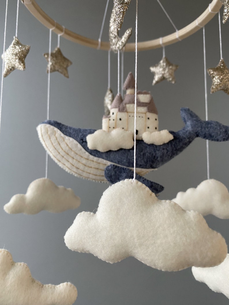 Puede incluir: Un m&oacute;vil para beb&eacute; con una ballena azul, nubes blancas y estrellas doradas con purpurina. La ballena lleva un peque&ntilde;o castillo marr&oacute;n.