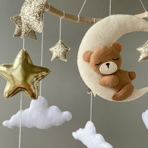 May include: A wooden baby mobile with a white felt crescent moon, a brown felt bear sleeping on the moon, and gold glitter stars.