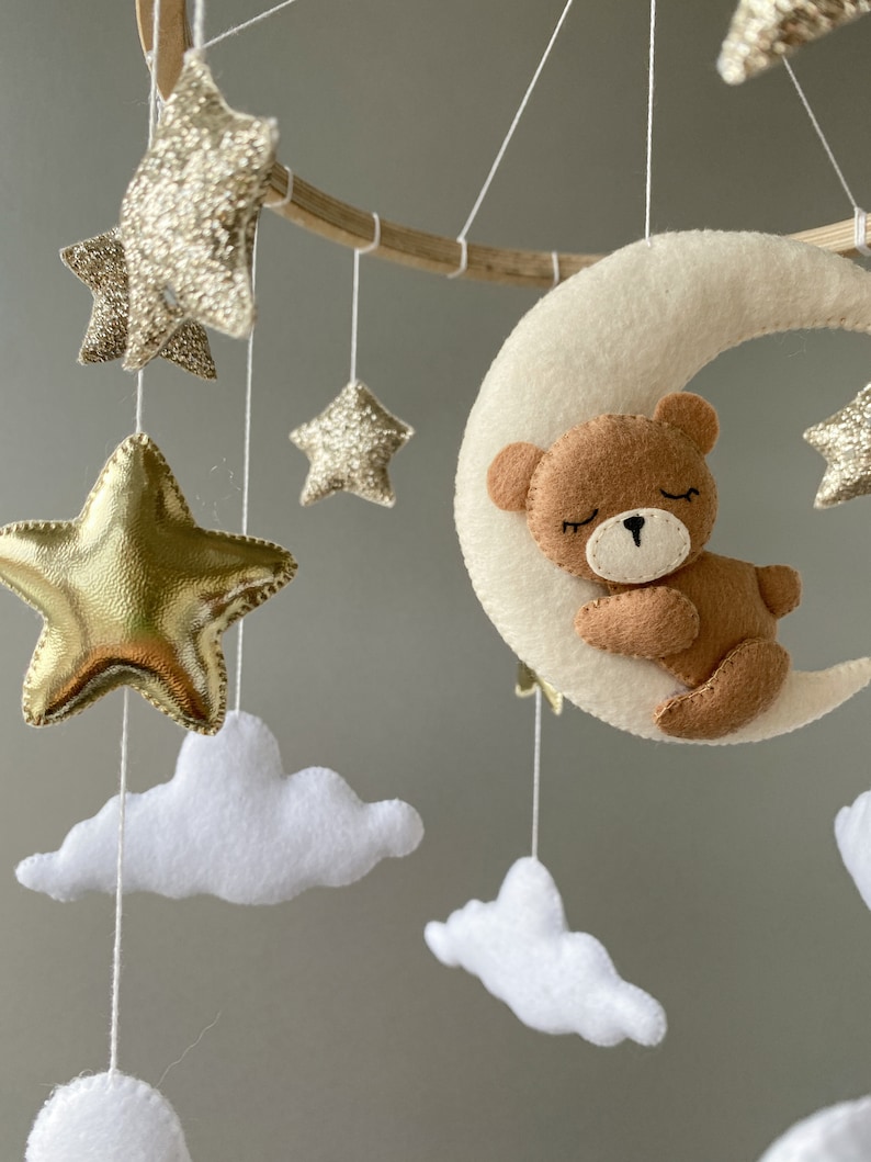 May include: A wooden baby mobile with a white felt crescent moon, a brown felt bear sleeping on the moon, and gold glitter stars.