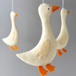 May include: A white felt goose mobile with orange felt feet and beaks. The geese are hanging from a wooden ring.