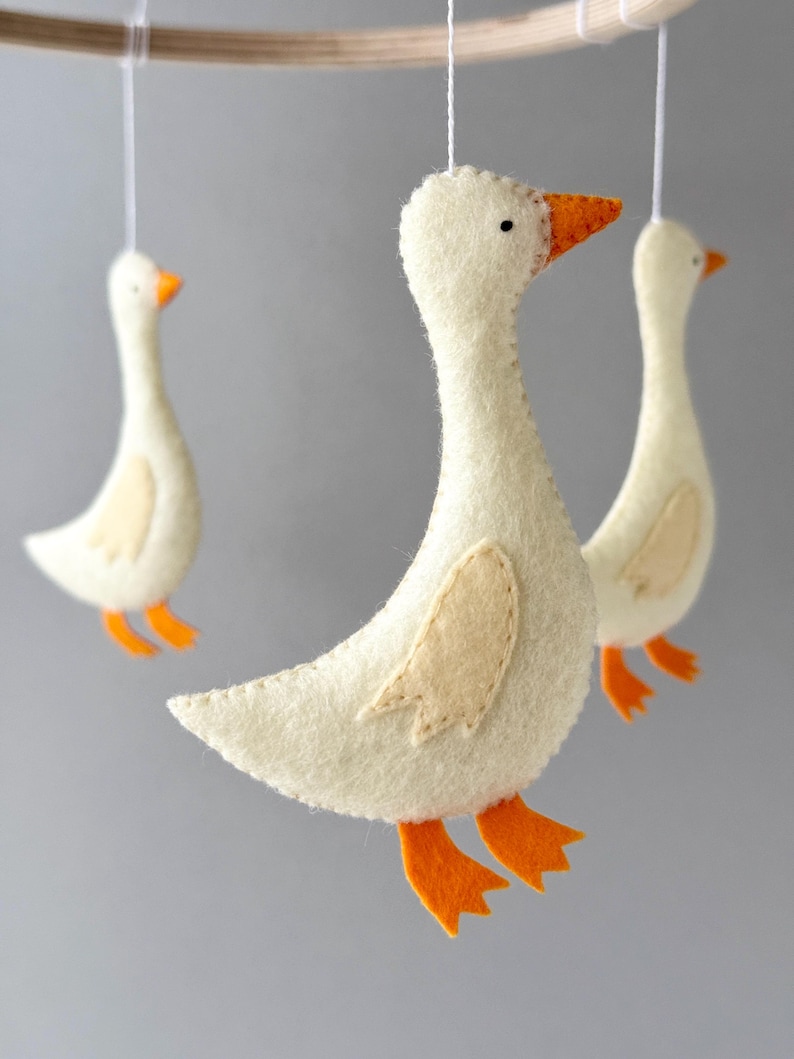 May include: A white felt goose mobile with orange felt feet and beaks. The geese are hanging from a wooden ring.