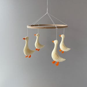 May include: A wooden mobile with four felt geese hanging from it. The geese are white with orange beaks and feet. The mobile is hanging from a white string.