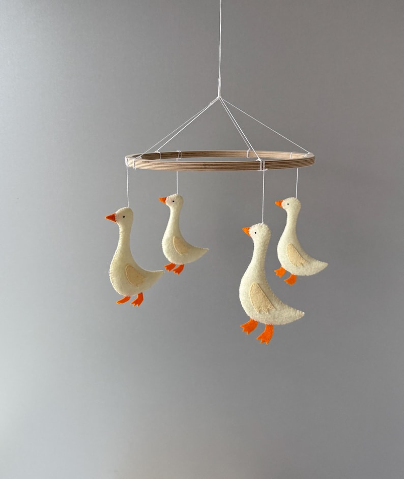 May include: A wooden mobile with four felt geese hanging from it. The geese are white with orange beaks and feet. The mobile is hanging from a white string.