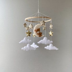 May include: A wooden baby mobile with a crescent moon, a teddy bear, and gold glitter stars hanging from white felt clouds.