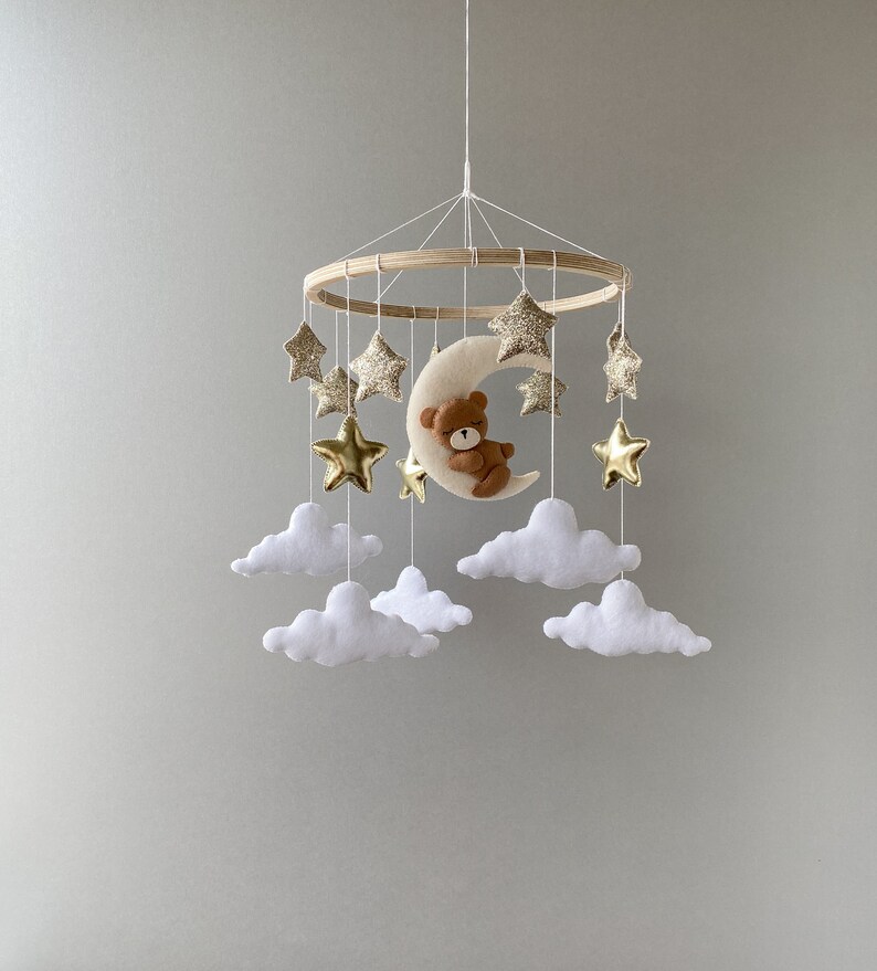 May include: A wooden baby mobile with a crescent moon, a teddy bear, and gold glitter stars hanging from white felt clouds.