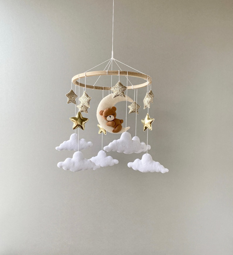 May include: A wooden baby mobile with a crescent moon, a teddy bear, and gold glitter stars hanging from white felt clouds.
