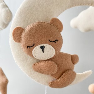 May include: A brown felt teddy bear with closed eyes is sleeping on a white felt crescent moon. The moon is part of a mobile with other felt clouds and stars.