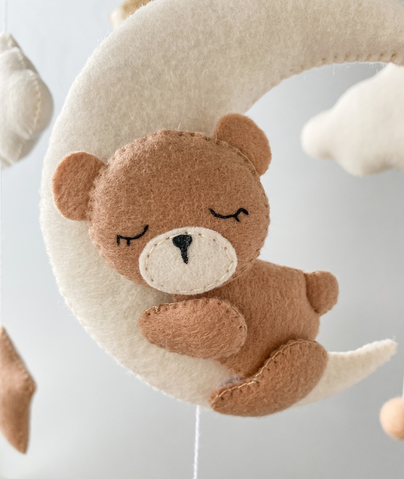 May include: A brown felt teddy bear with closed eyes is sleeping on a white felt crescent moon. The moon is part of a mobile with other felt clouds and stars.