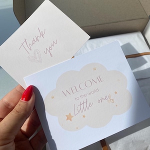 May include: A white card with the text "Welcome to the world little one" in a cloud shape with gold stars. The card is on top of a brown box with a white tissue paper lining. A thank you card with a pink heart is visible in the background.