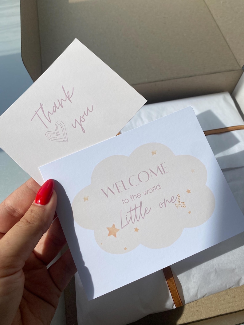May include: A white card with the text "Welcome to the world little one" in a cloud shape with gold stars. The card is on top of a brown box with a white tissue paper lining. A thank you card with a pink heart is visible in the background.