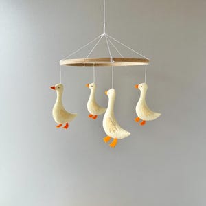 May include: A wooden mobile with four felt geese hanging from it. The geese are white with orange beaks and feet. The mobile is suspended from a white string.