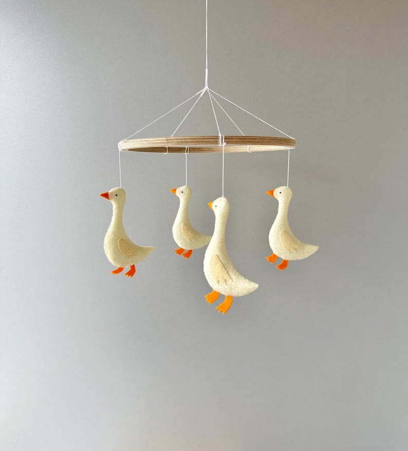 May include: A wooden mobile with four felt geese hanging from it. The geese are white with orange beaks and feet. The mobile is suspended from a white string.