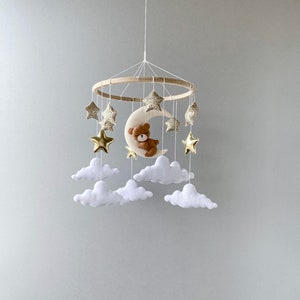 May include: A wooden baby mobile with a crescent moon, stars, and clouds. The moon has a brown teddy bear sleeping on it. The stars are gold with glitter. The clouds are white.