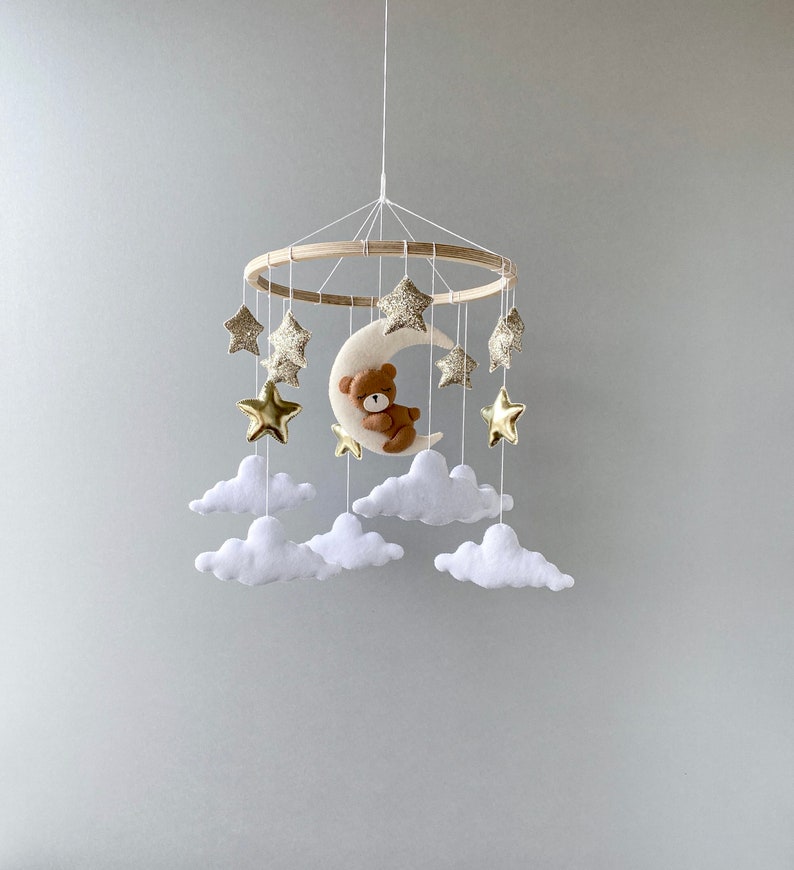 May include: A wooden baby mobile with a crescent moon, stars, and clouds. The moon has a brown teddy bear sleeping on it. The stars are gold with glitter. The clouds are white.