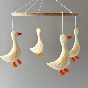 May include: A white felt goose mobile with four geese hanging from a wooden ring. The geese have orange beaks and feet.