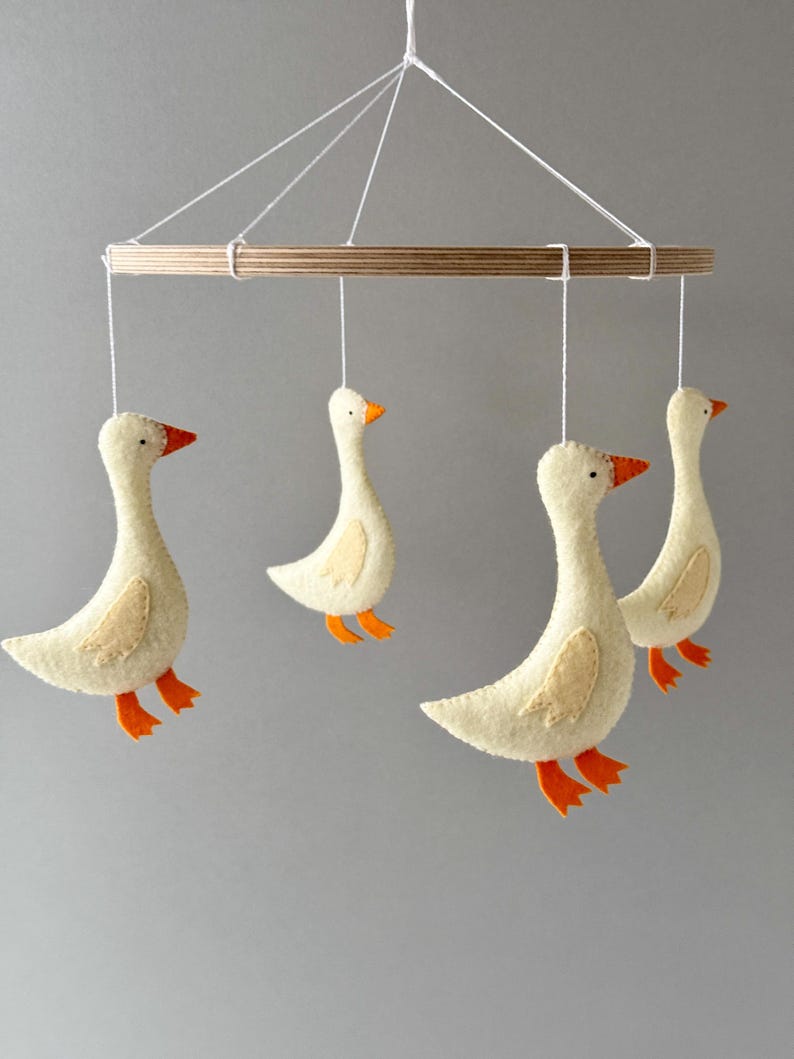 May include: A white felt goose mobile with four geese hanging from a wooden ring. The geese have orange beaks and feet.
