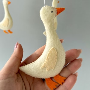 May include: A white felt goose with orange beak and feet, hanging from a mobile. The goose is handmade and has a simple design.