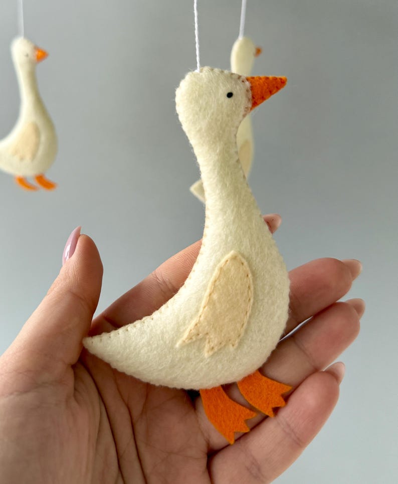 May include: A white felt goose with orange beak and feet, hanging from a mobile. The goose is handmade and has a simple design.