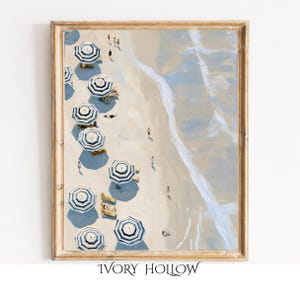 May include: A framed art print featuring an aerial view of a sandy beach with multiple blue and white striped umbrellas, small figures of people, and light blue ocean waves washing onto the shore, all enclosed in a light wood frame.