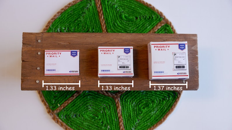 May include: Three small white cardboard boxes labeled "PRIORITY MAIL" with red and blue accents, arranged on a wooden plank. The boxes measure approximately 1.33 inches and 1.37 inches wide. The wooden plank is mounted on a green and brown woven background.