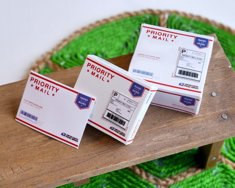 May include: Three white Priority Mail boxes and an envelope with red and blue accents. The boxes and envelope have the words "Priority Mail" and "Tracked & Insured" printed on them. The boxes are on a wooden surface.