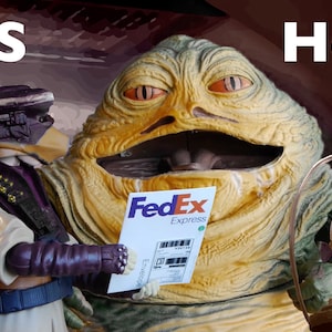 以下が含まれることがあります： 緑と黄色のジャバ・ザ・ハットのフィギュアが、FedEx Expressの配送ラベルを持っています。フィギュアは笑顔で、大きな口をしています。紫と白のアクションフィギュアがジャバの隣に立っていて、ラベルを持っています。テキスト「IT'S Here!」は、茶色の背景に白い文字で書かれています。