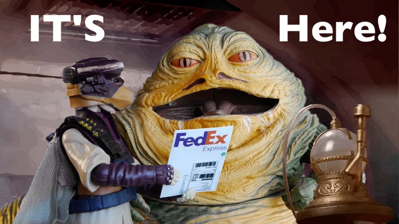 以下が含まれることがあります： 緑と黄色のジャバ・ザ・ハットのフィギュアが、FedEx Expressの配送ラベルを持っています。フィギュアは笑顔で、大きな口をしています。紫と白のアクションフィギュアがジャバの隣に立っていて、ラベルを持っています。テキスト「IT'S Here!」は、茶色の背景に白い文字で書かれています。